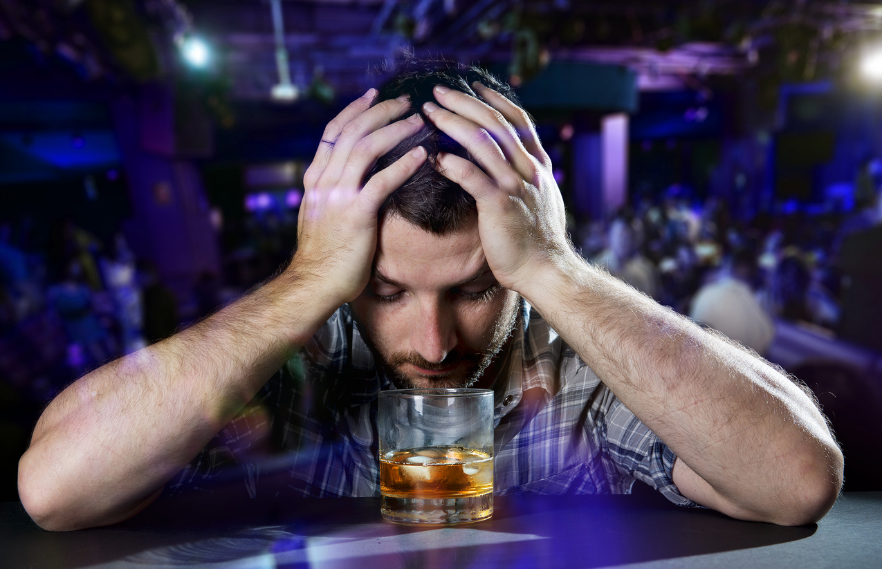 young drunk man in casual shirt depressed and wasted with hands on his head drinking a glass of whiskey at evening disco nightclub feeling dejected and overwhelmed by problems at work or at home